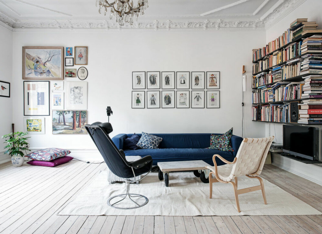 Vitt vardagsrum med stuckatur, tavelvägg och bokhylla. Blås soffa och ljust trägolv. 