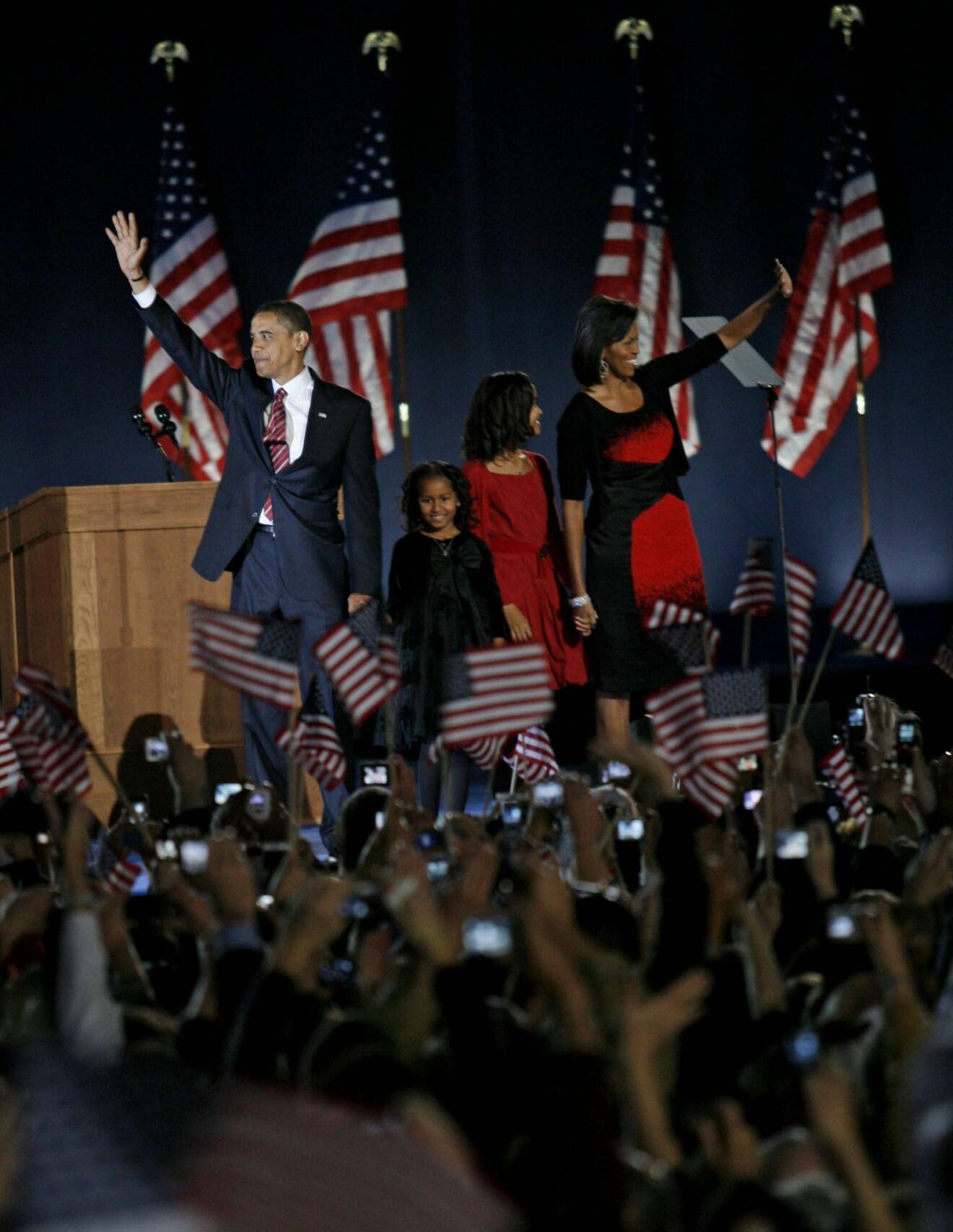 Familjen Obama står på en scen och vinkar till folkmassorna som viftar med amerikanska flaggor