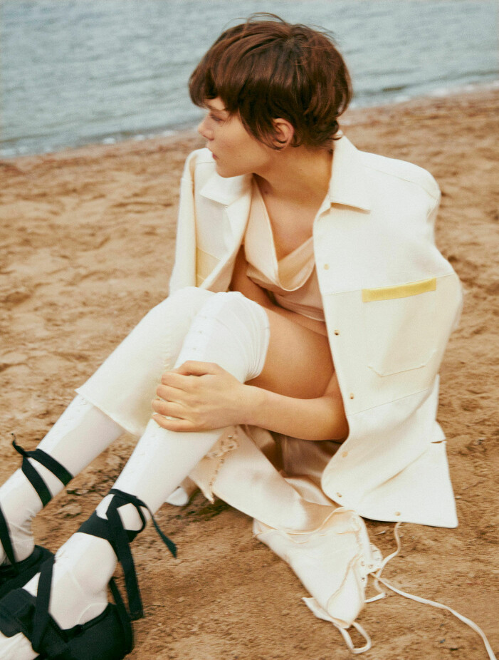 Klänning, 10 000 kr, Jade Cropper. Skjorta, 5 190 kr, Max Mara. Strumpor 3 700 kr, Acne Studios. Skor, pris vid beställning, Sportmax. Modellen sitter på en strand och hon har på sig en vit klänning från Jade Cropper och en skjorta över axlarna från Max Mara