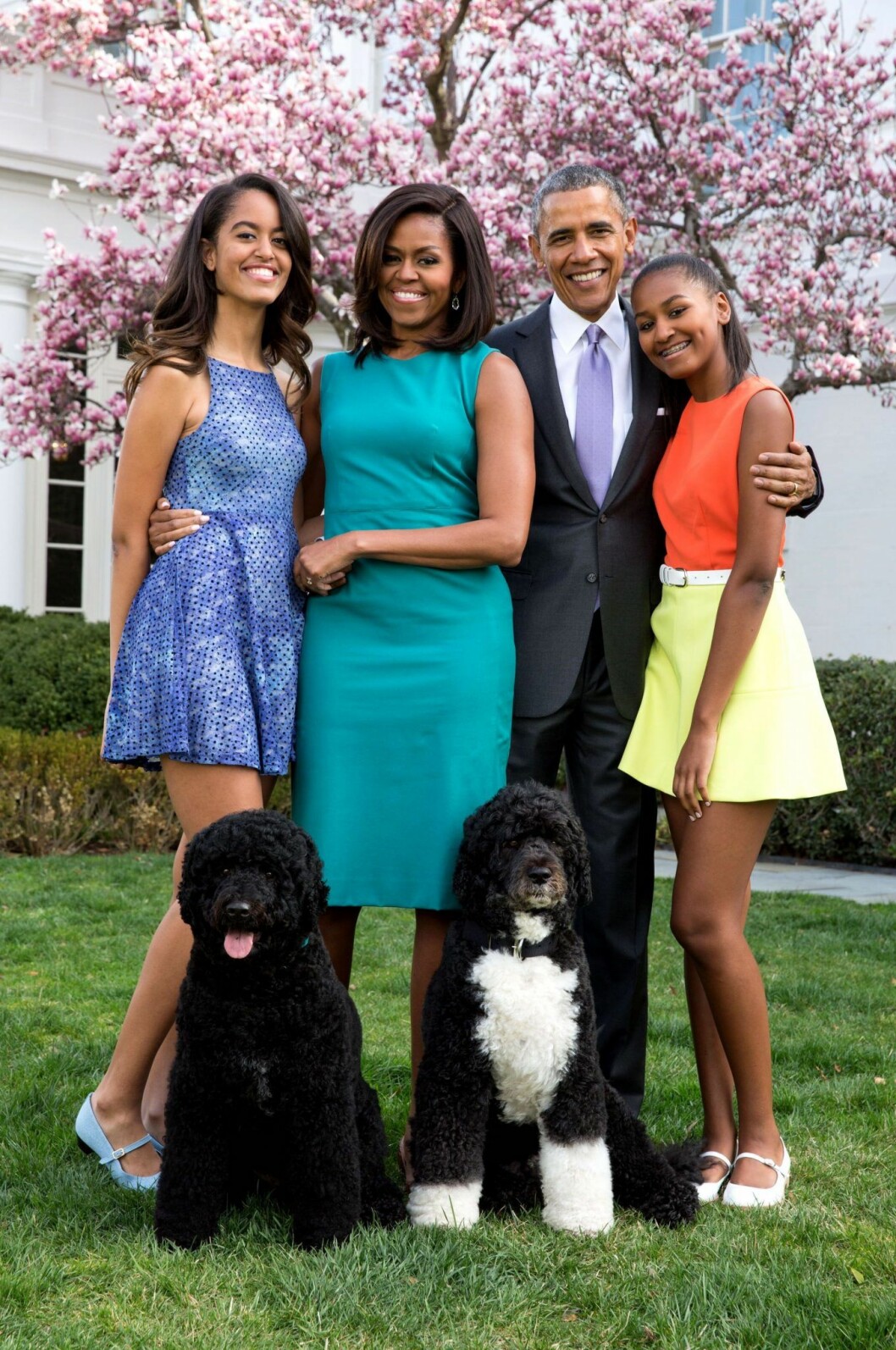 Här har döttrarna hunnit bli ännu större och året är 2015. Malia i blå klänning är 17 år och Sasha i gult och orange är 14 år. Michelle, Malia, Barack och Sasha Obama. står i trädgården i färgglada kläder och med två hundar framför sig