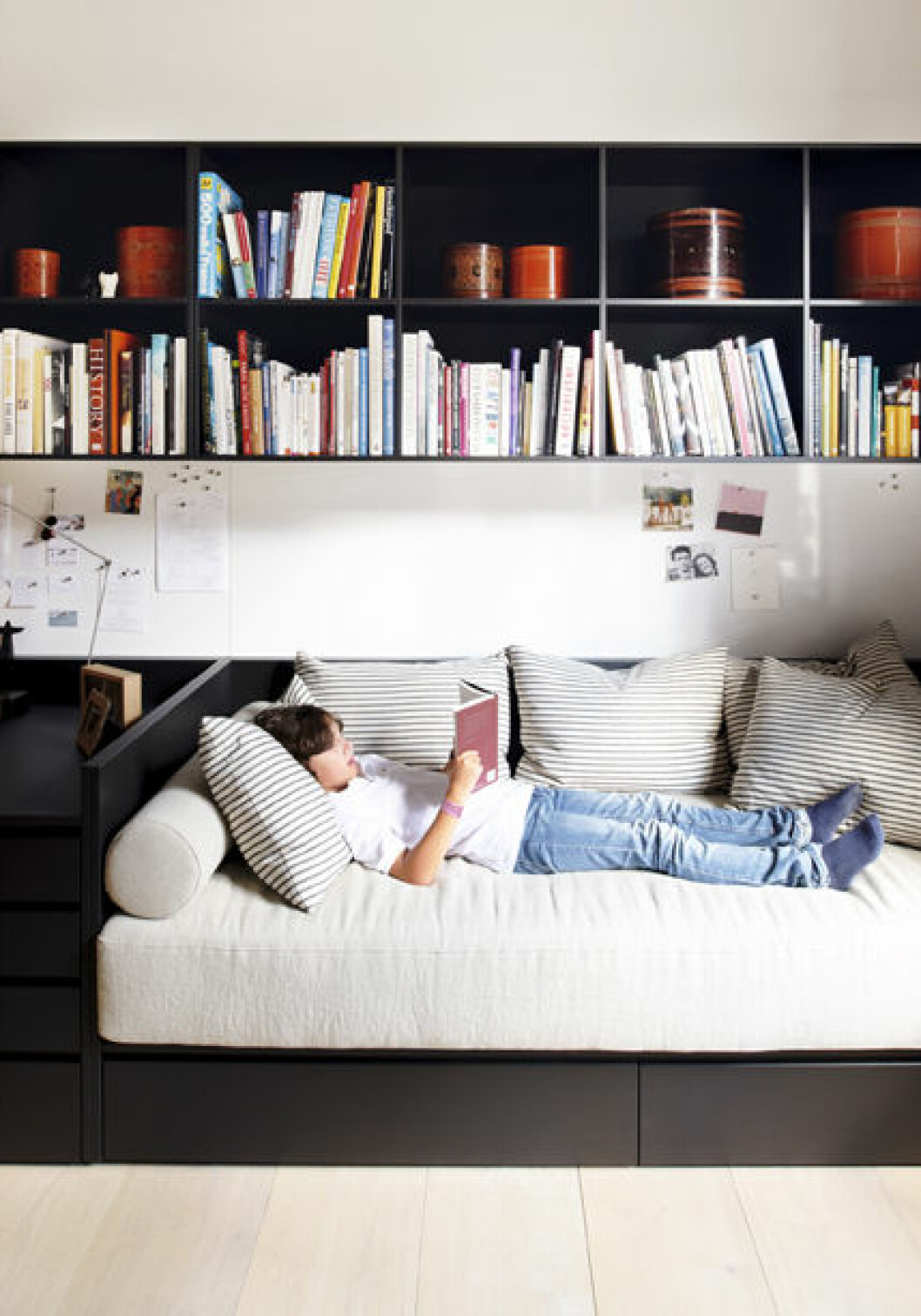 Filippa Knutssons son läser en bok i hemma i soffan med bokhylla ovanför.