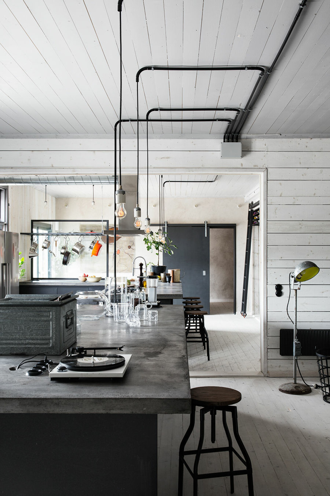 I det rymliga köket finns gott om plats att umgås och laga mat tillsammans. De stora bänkskivorna i betong är gjutna på plats. Barpallar Swansea från Barstol.se. Kök, Deer Design/Johan Israelson. Lampsocklar av industrikeramik med klara glödlampor. Skjutdörr, design Johan Israelson/Norra Gotland. Konstverk på den bortre väggen av Kinga Fågelberg. Stege och vinställ, Johan Israelson/Norra Gotland. Industrigolvlampa, secondhand. Köksö i grå granit. Heltäckande vitt trärum
