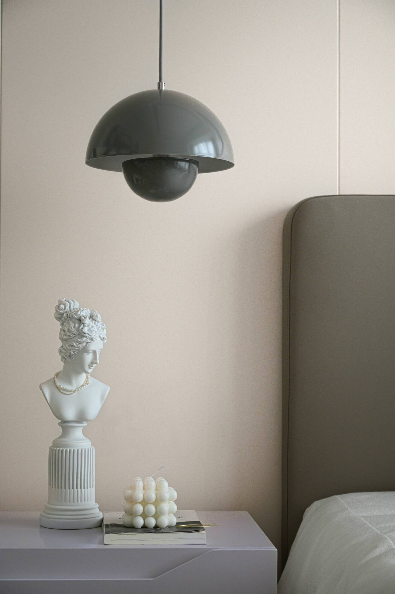 Hängande grå taklampa ovanför en brun sänggavel, en vit staty och ett bubbligt ljus