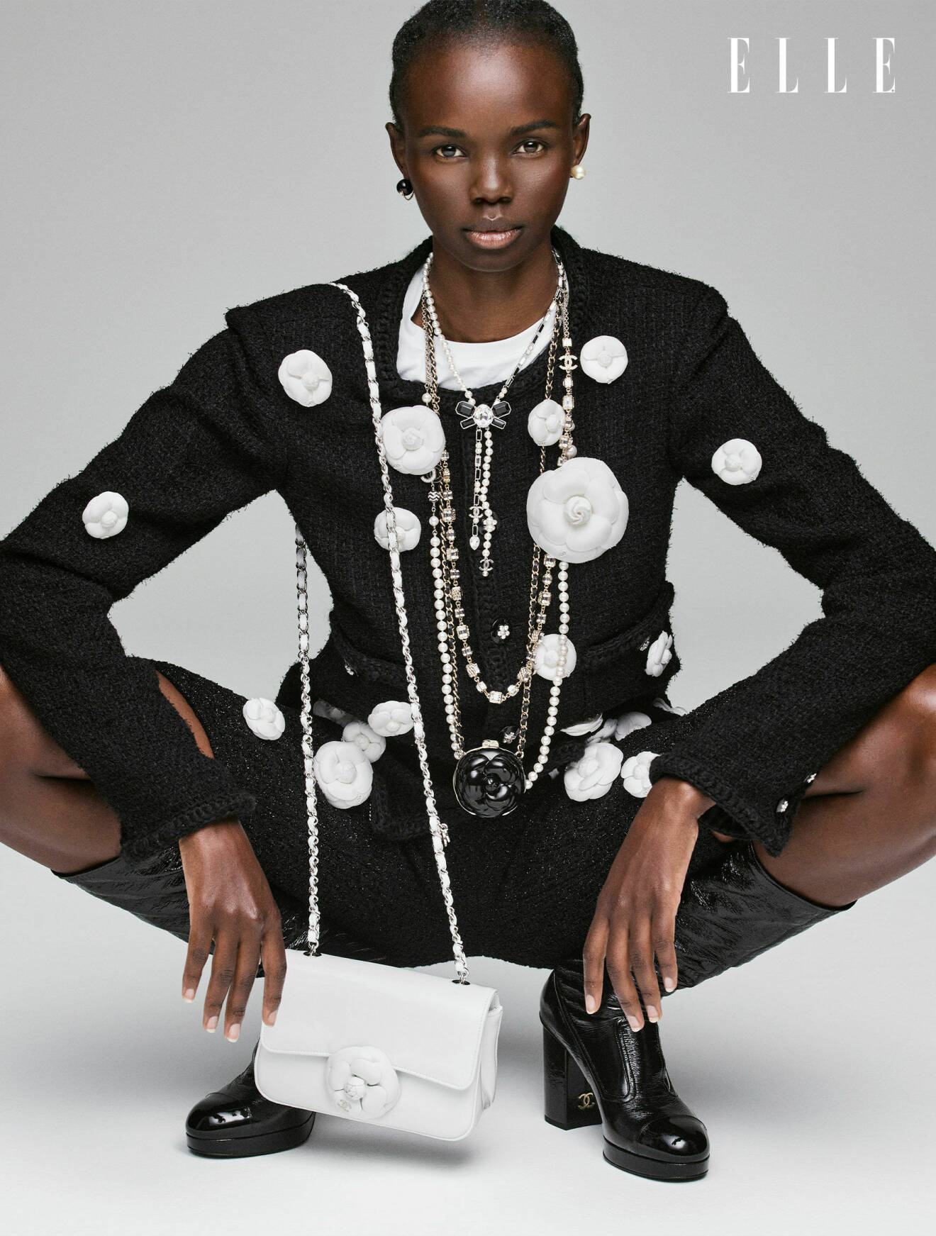 Modellen sitter på huk och hon har på sig en svart kavaj med vita blommor, svarta shorts, en vit miniväska och flera olika halsband.