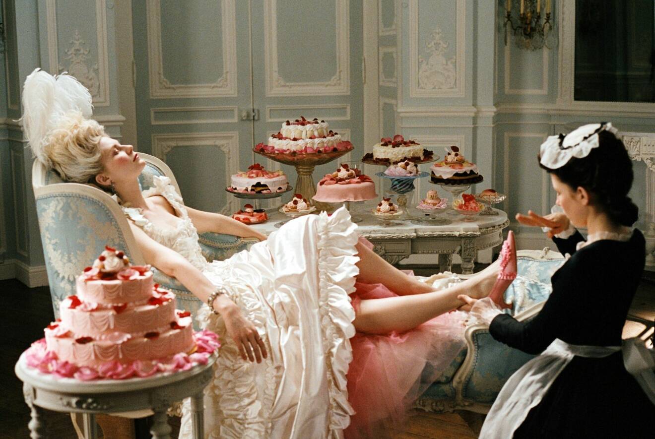 Kirsten Dunst i rolle som Marie Antoinette i vit klänning och rosa klackar, omgiven av bakelser.