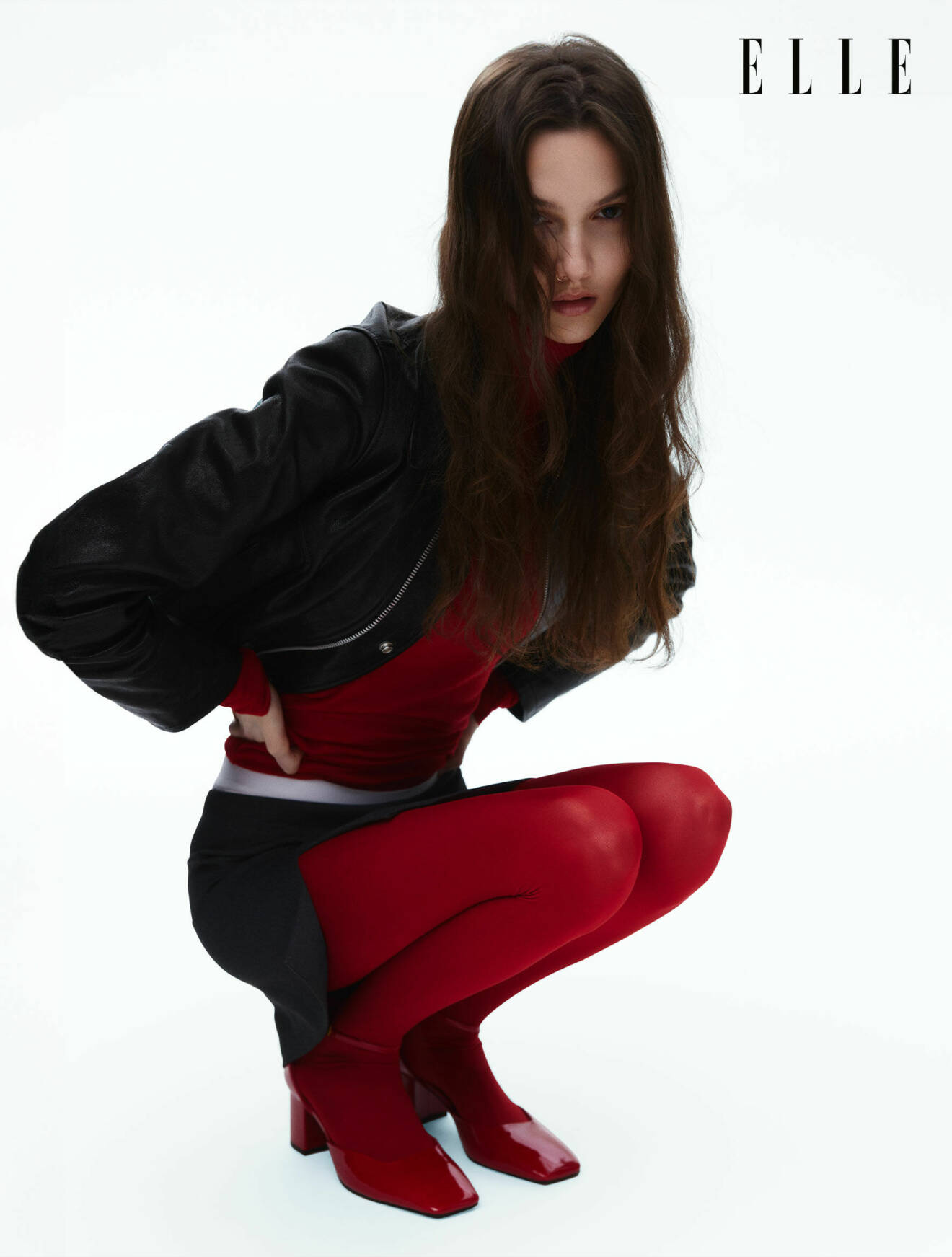 Modellen Alma sitter på huk, hon har på sig en svart jacka, röd polotröja, svart kjol och röda strumpbyxor