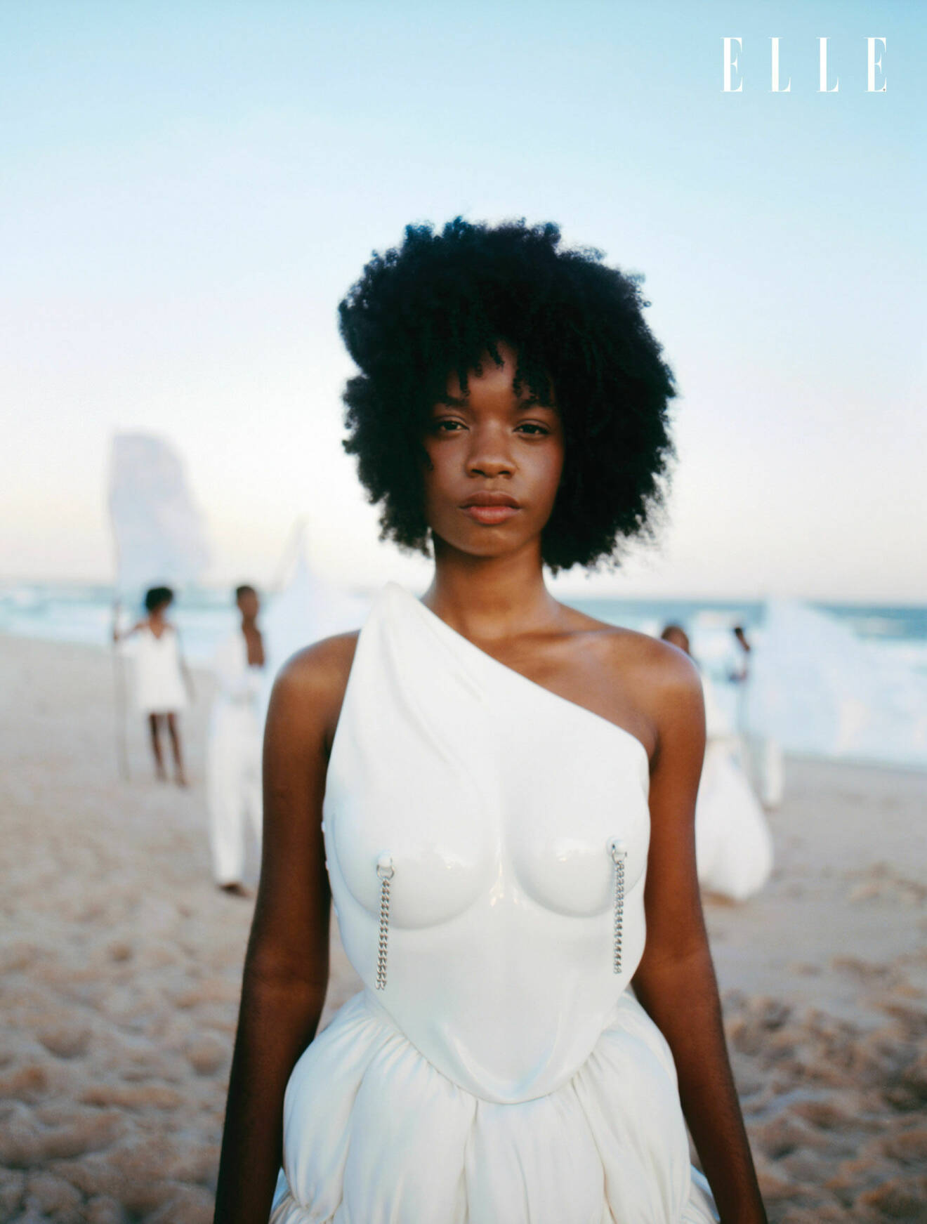 Modellen står på en strand, hon är klädd i en vit topp från Artemisi och en vit kjol från Handred