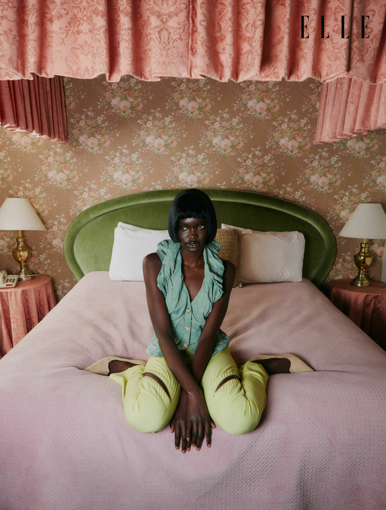 Fotomodellen sitter i en säng, hon har på sig en grön topp och gula byxor