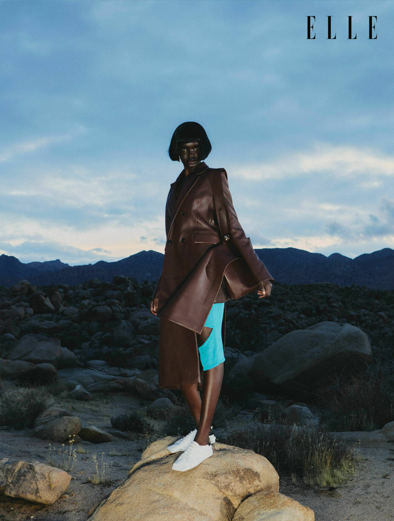 Fotomodellen poserar mot ett öppet landskap, hon har på sig en brun kappa och turkosa byxor