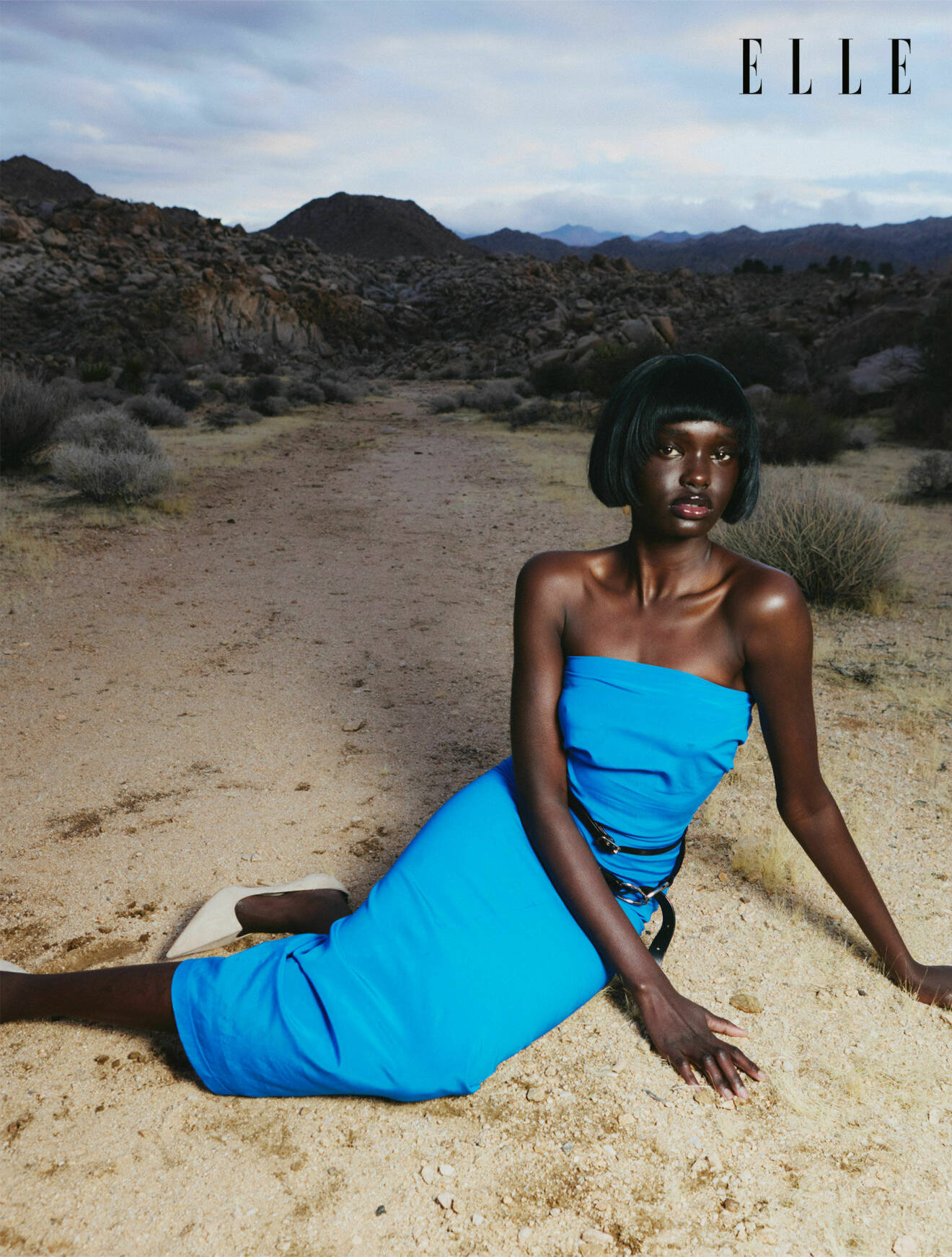 Fotomodellen sitter på marken, hon har på sig en blå ärmlös klänning och vita pumps