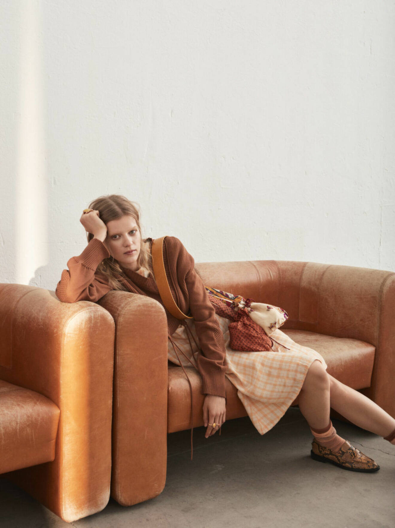Modell halvsittandes i rostbrun soffa, iklädd klänning från Acne Studios.