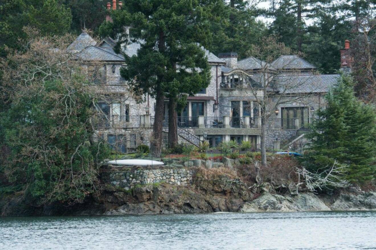Ett stort grått hus i sten som ligger dolt bland träd och med vatten framför