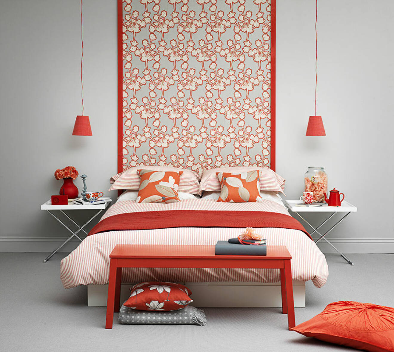 Modern grey and coral bedroom. Grey painted wall with strip of bold grey and coral floral wallpaper framed by coral lamp shades hanging from long cords, white folding bedside tray tables, striped coral and white bedding, red bench style table at tip of bed, grey carpet on floor, IH 05/2012 Pub Orig