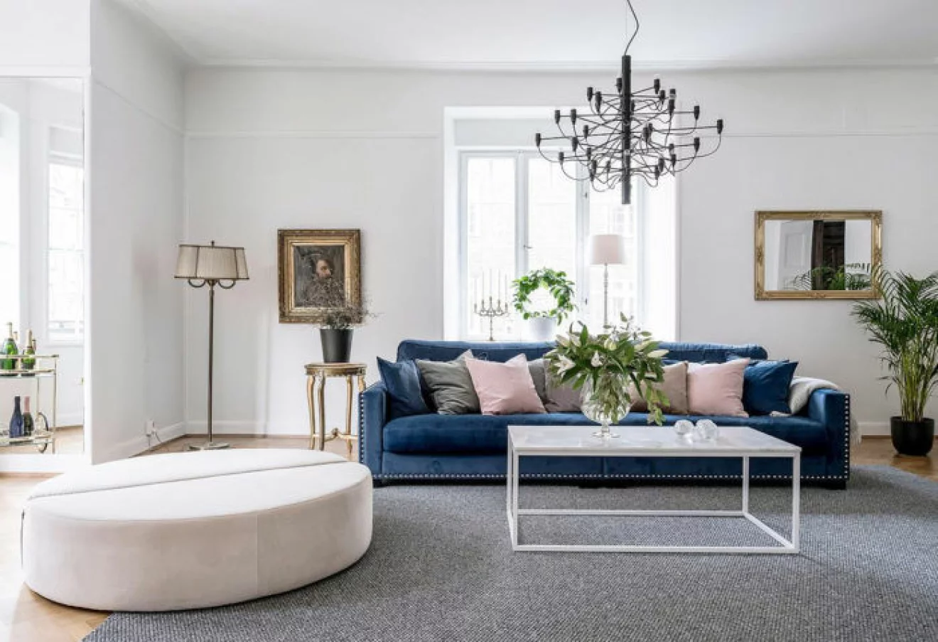 Vardagsrum med stor dekorativ lampa, vitt stilrent soffbord och en blå soffa i sammet. En rosa tillhörande puff.
