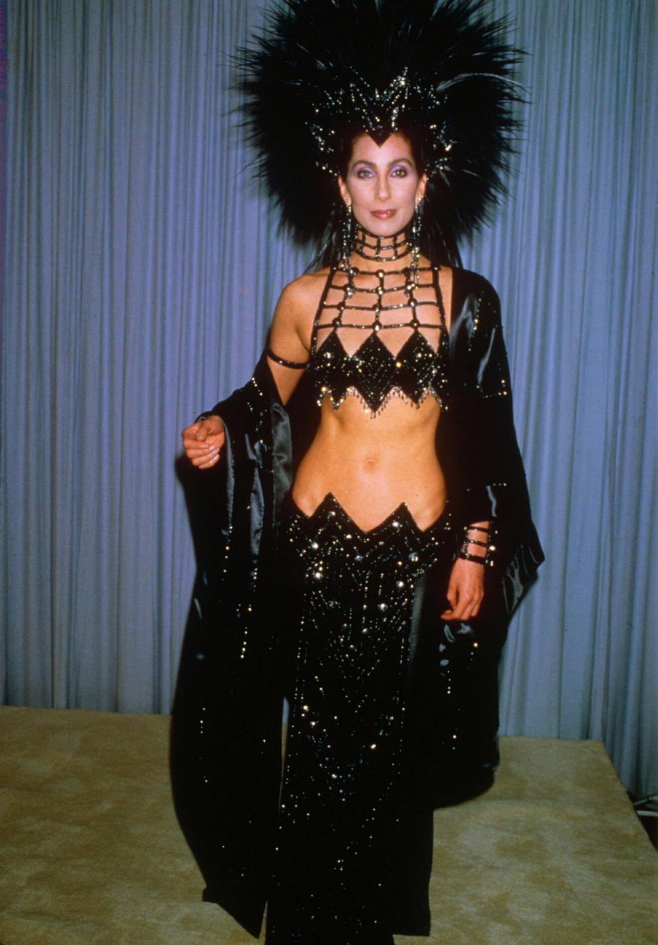 Cher i en svart glittrig showgirl kreation med en stor fjäderdekoration i håret.