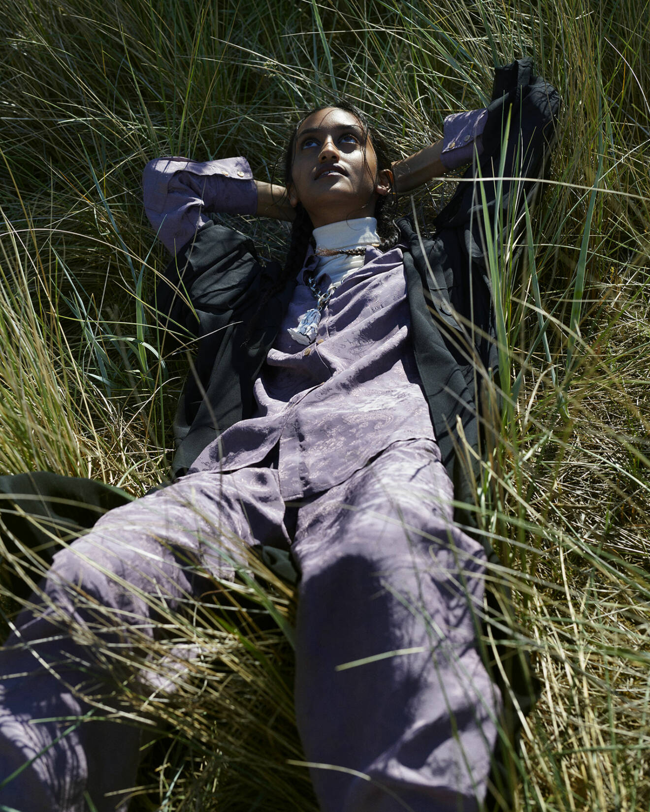 Modellen ligger i gräset och har på sig en svart skjortklänning, lila skjorta med matchande byxor och en vit polotröja under