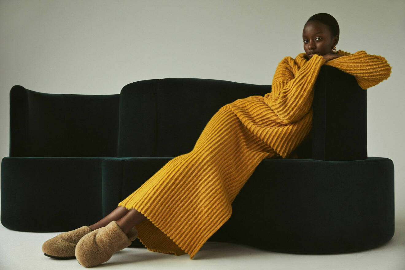 Modellen sitter i en modulsoffa och har på sig en gul, stickad tröja och kjol