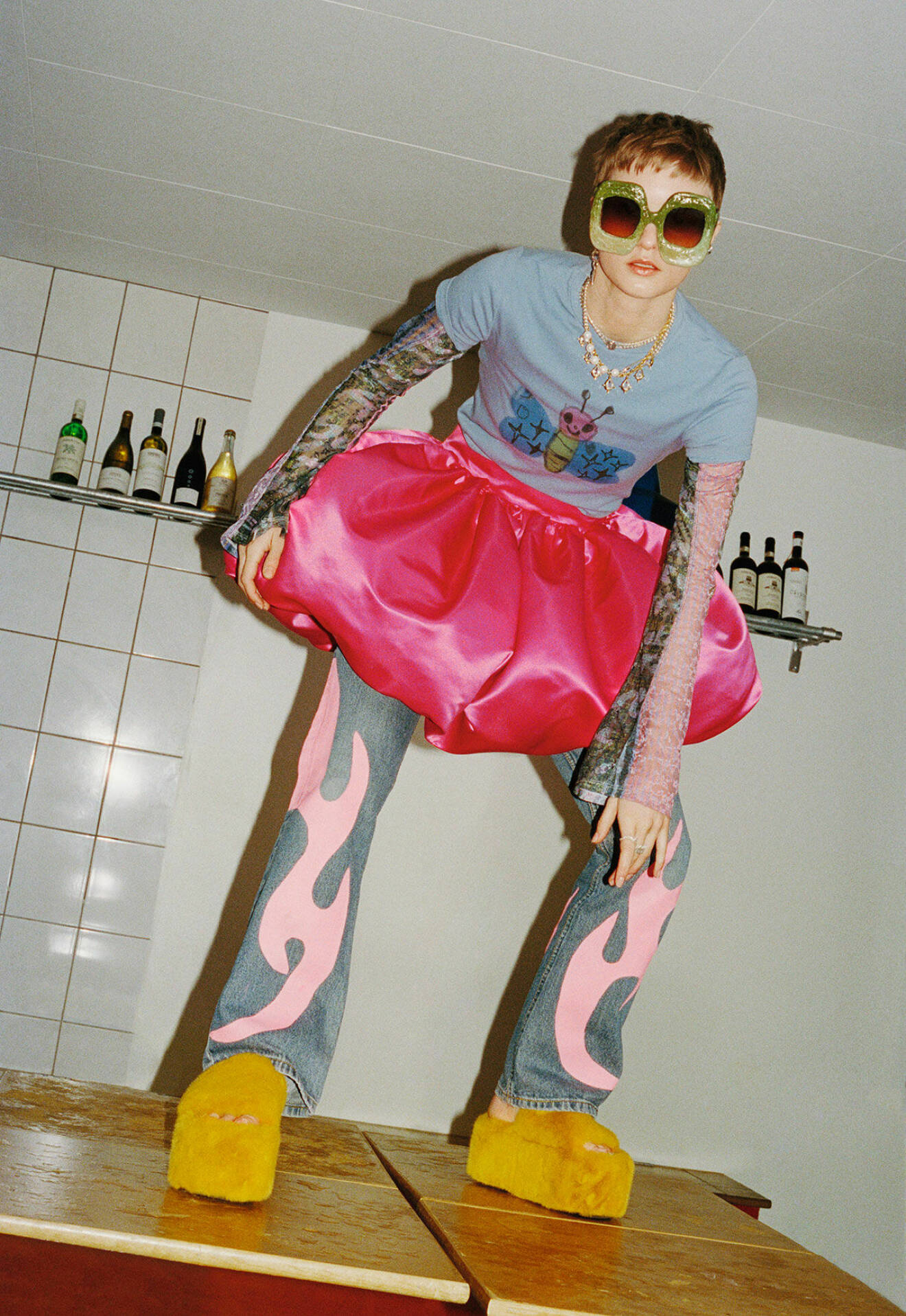 Modellen står på ett bord, hon har på sig en rosa kjol, mönstrade jeans och en t-shirt med tryck
