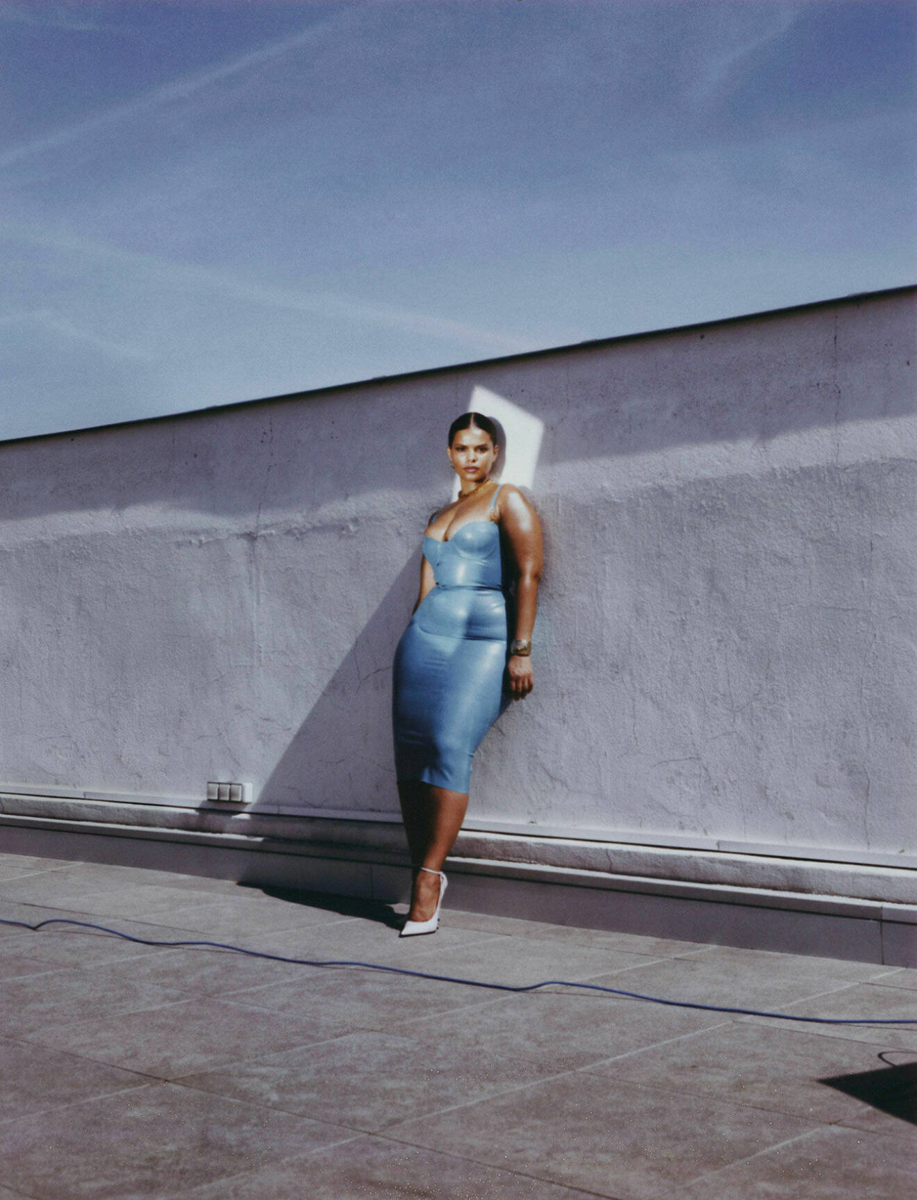 Modellen står lutad mot en vägg, hon har på sig en blå klänning och vita pumps