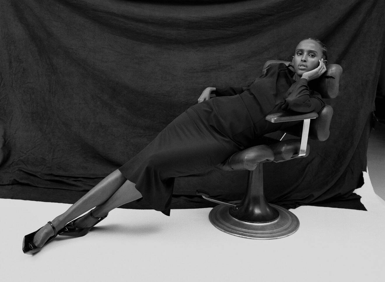 Fotomodellen halvligger ner i en stol och har på sig en svart klänning och svarta skor, allt från Prada.