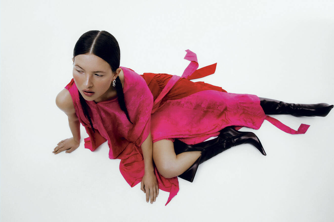 Fotomodellen sitter på golvet, hon har på sig en ceriserosa klänning och svarta boots.