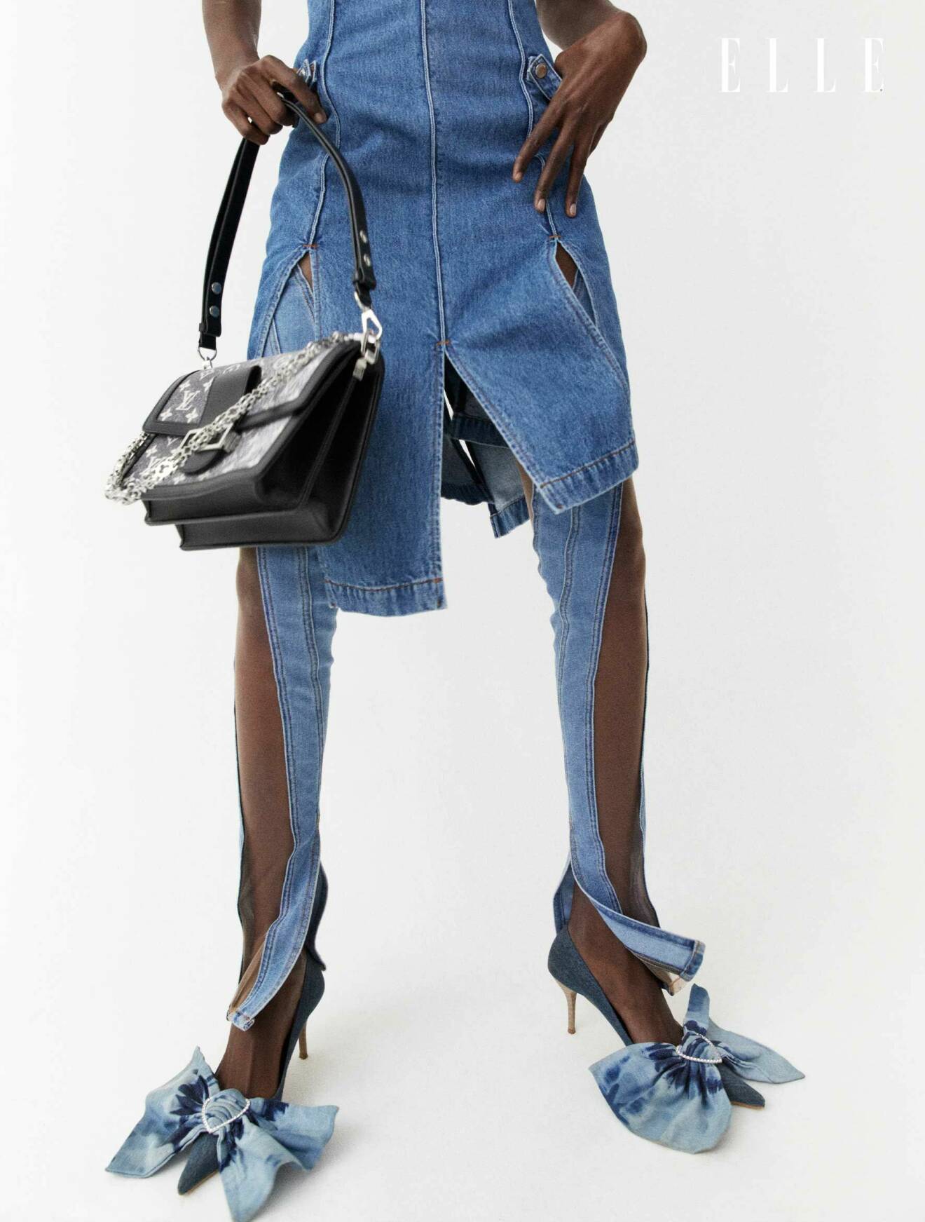 Fotomodellen bär en klänning i denim tillsammans med en svart handväska och skor med denimrosetter.