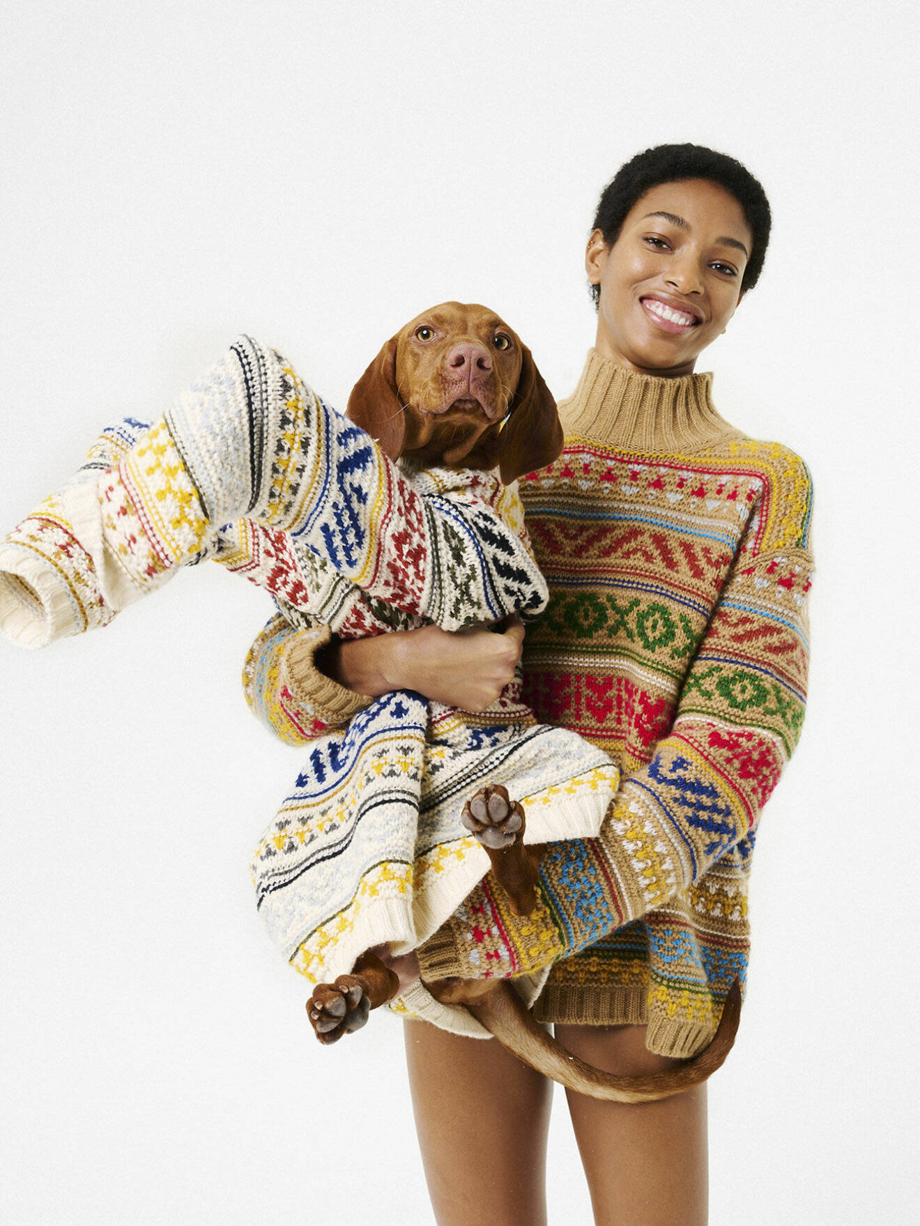 Fotomodellen har på sig en stickad polotröja med mönster och bär en hund som även den har på sig en stickad tröja med mönster, båda från Loro Piana.