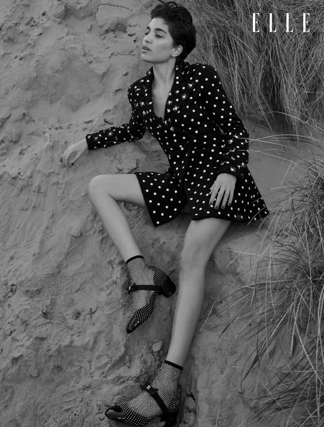 Modellen sitter i sanden, hon bär en svart kavajklänning med vita prickar från CHanel.