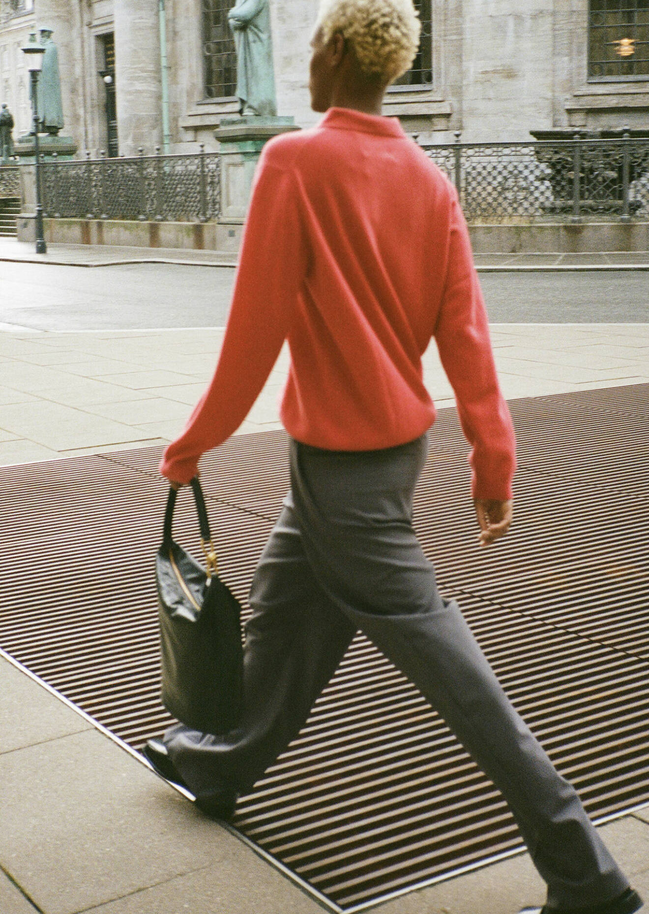 Fotomodellen har på sig en röd tröja, grå byxor och en svart handväska.
