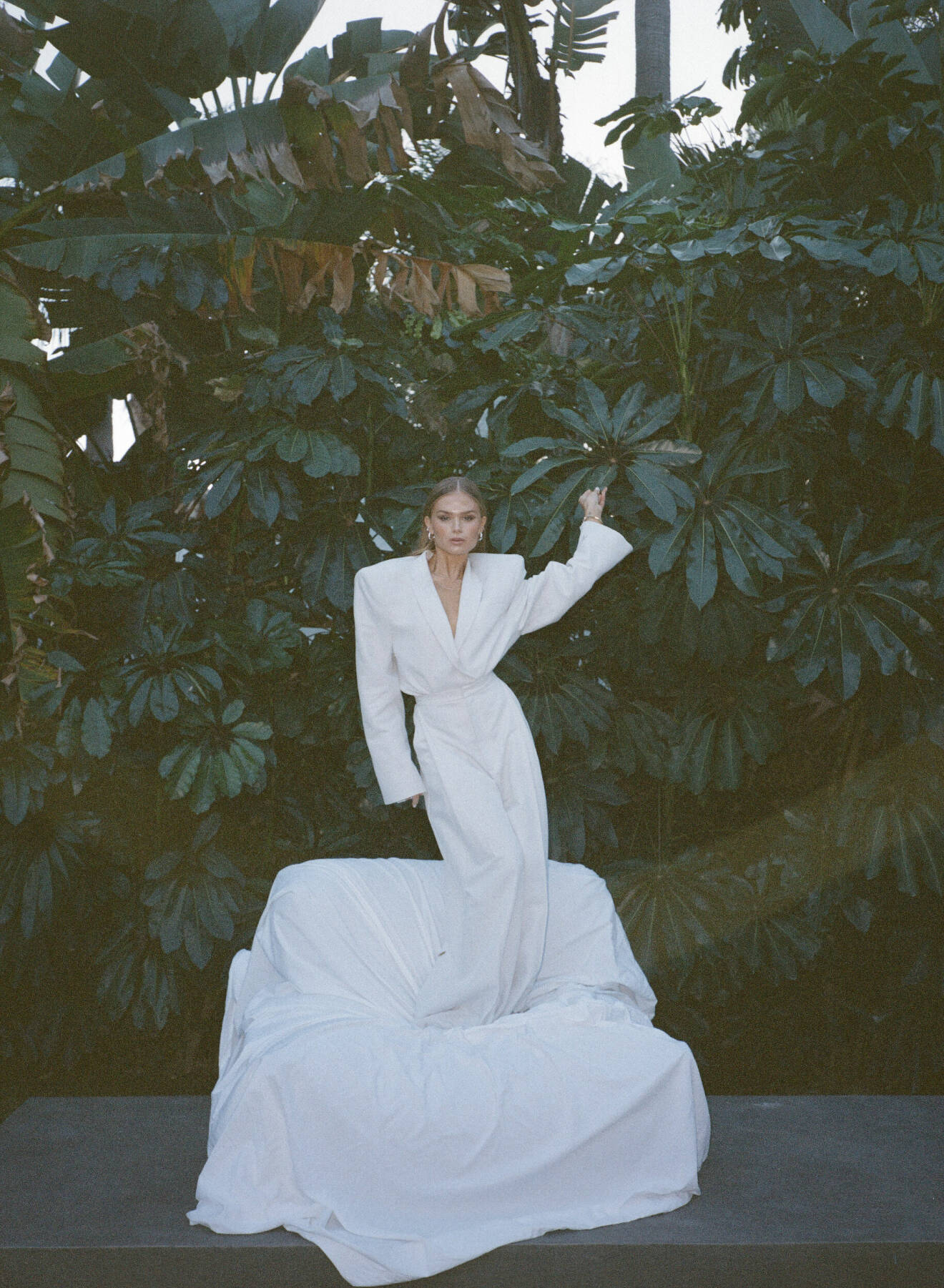 Hanna Schönberg bland grönska på, i en vit blazer och kostymbyxor. Ståendes på en soffa, täckt med ett vitt lakan.