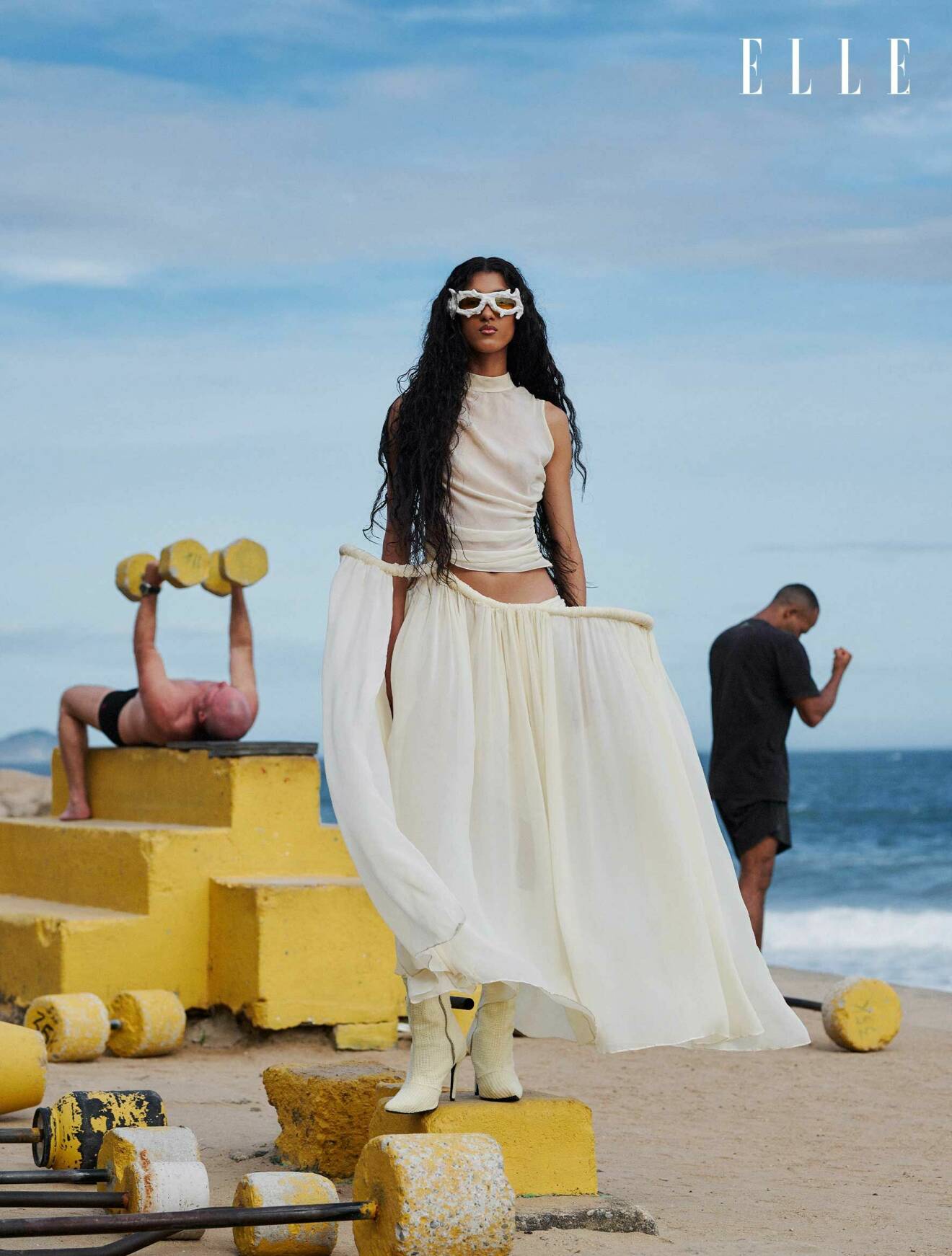 Modellen står på stranden klädd i en lång vit klänning från Lucas Leão