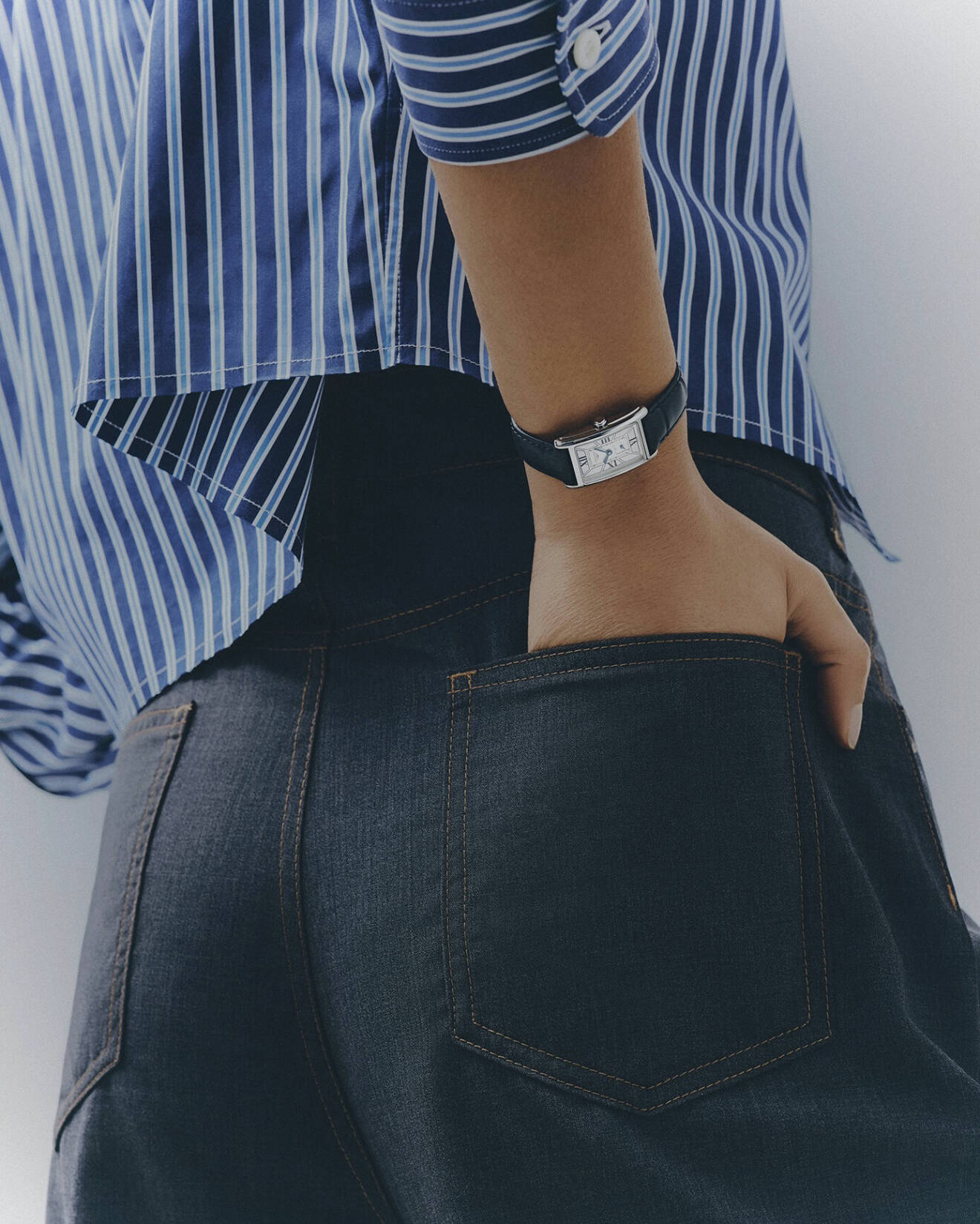 Modellen har på sig en randig skjorta i blått och jeans, båda från Miu Miu/Mytheresa. Hon har handen i backfickan och bär en klocka från Longines.