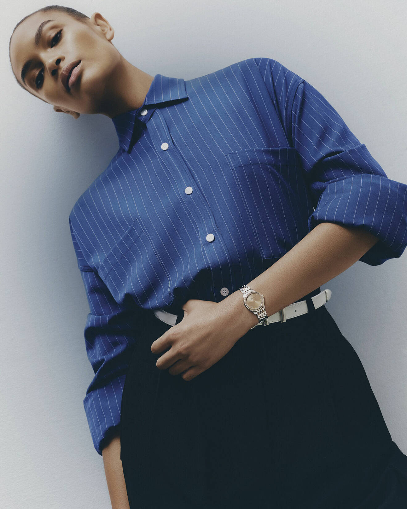 Modellen bär en randig blå skjorta och svarta byxor från The Frankie Shop. På armen har hon en klocka från Omega.