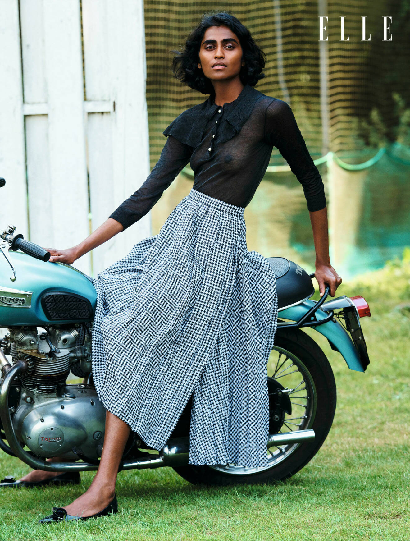 Modellen sitter på en motorcykel, hon har på sig en svart blus och lång smårutig kjol, allt från Dior.