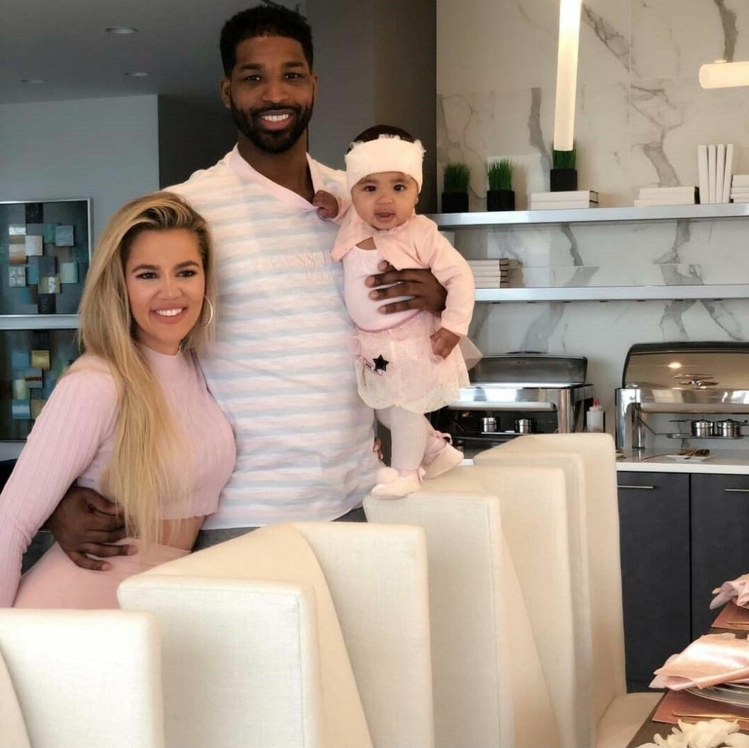 Khloé Kardashian och Tristan Thompson står bakom några stolar vid ett köksbord. Tristan håller en arm runt Khloé och håller dottern True med den andra