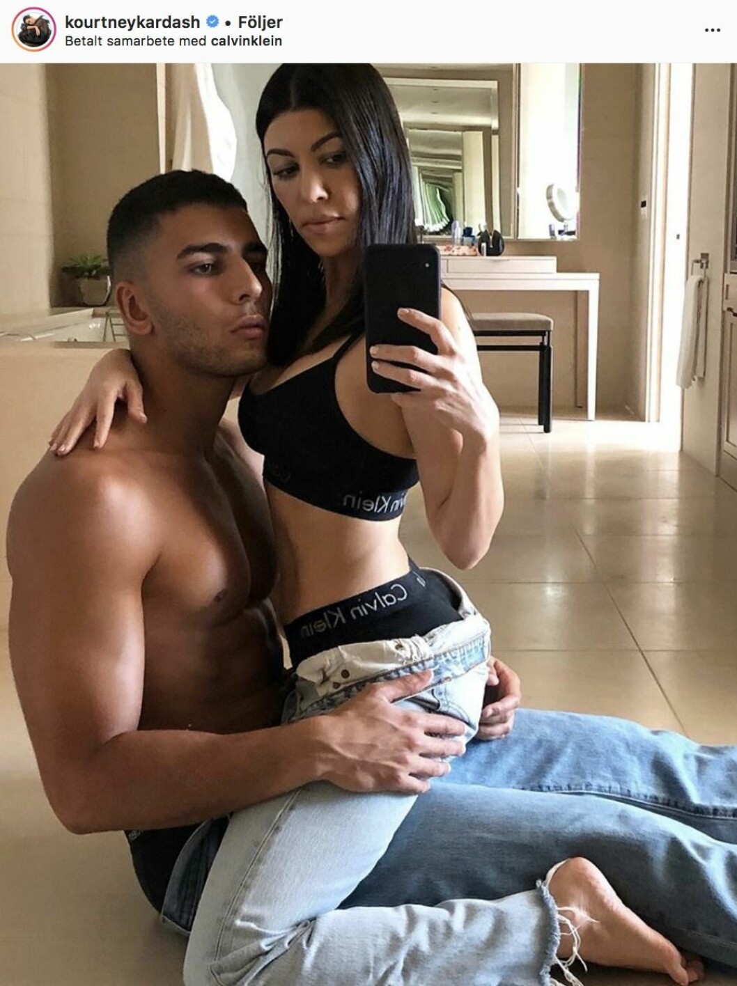 Kourtney Kardashian sitter i Younes Bendjimas knä på golvet. Han har bara jeans på sig och hon har jeans och en sporttopp