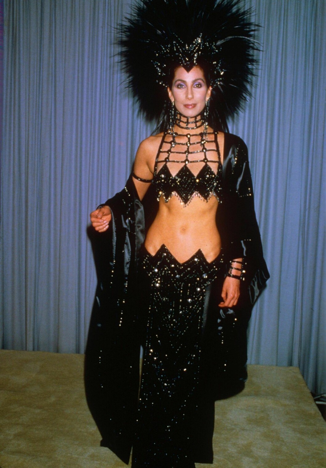 Cher manifesterar i en riktig showgirl outfit. Cher i en svart glittrig showgirl kreation med en stor fjäderdekoration i håret.