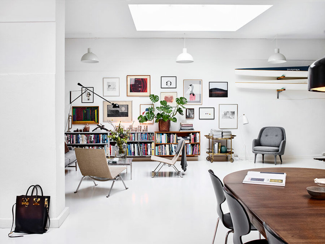 Allrummets bortre vägg är möblerad med Mogen Kochs bokhyllor och väggen ovan är tapetserad med bilder av baland andra Rasmus Nellemann, Simon Lindhardt och Cathrine Raben Davidsen. Vägglampan 265, i design Paolo Rizzato är med sitt industriella uttryck ett perfekt val till soffgruppen med PK22-stolar i canvas och PK61 soffbord i marmor, från Fritz Hansen. Modulljusstaken av Fritz Nagel var en födelsedagspresent. Jaime Hayon är designern bakom den grå länsstolen från Fritz Hansen. Här står den under parets vägghängda kajaker och vid sidan av Louises farmors gamla barbord som i nytolkning blivit til ett växtbord. I den stora hyllan har Anders sina fotoprylar. Den vita golvlampan är Grasshopper från Gubi, design Greta Magnusson Grossman.