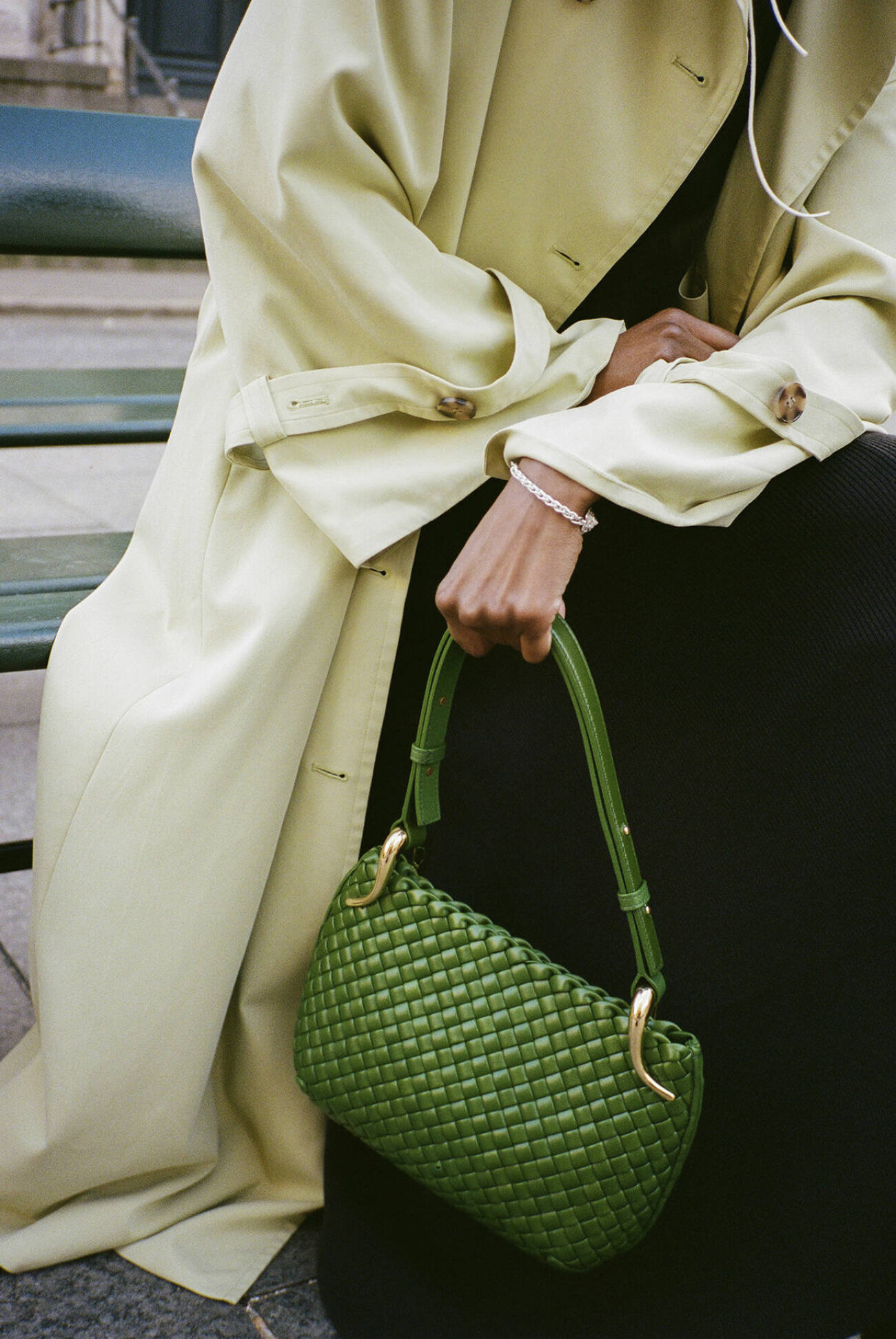 Modellen bär en beige trenchcoat, en svart klänning och bär en grön handväska.